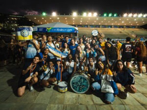 Grande grupo de integrantes da delegação da Atlética Finanças UFF reúne-se para uma foto noturna ao ar livre, celebrando o resultado da competição. Ao centro da imagem, estão expostos troféus conquistados pela equipe, enquanto os estudantes fazem gestos de comemoração e sorriem para a câmera.