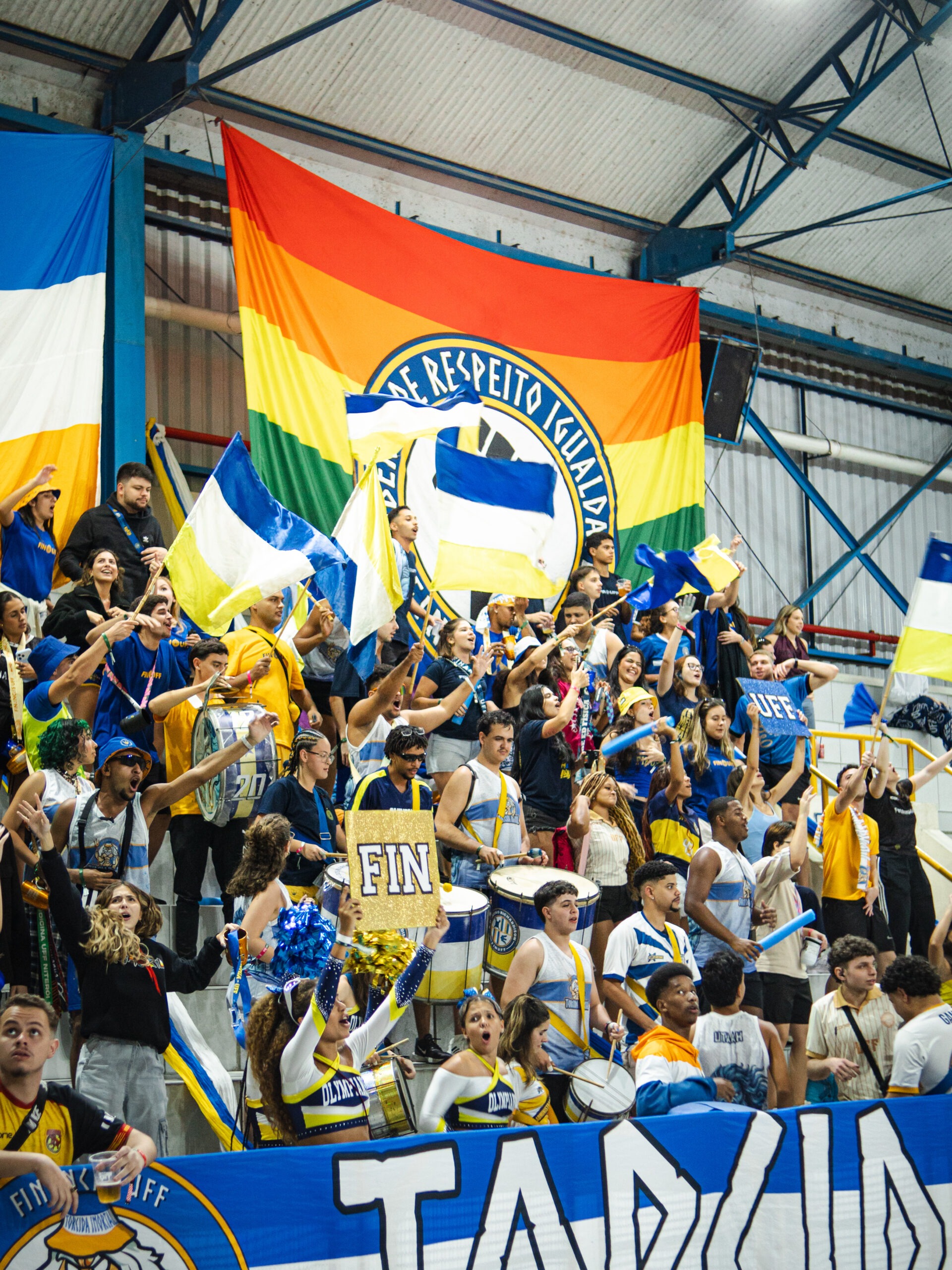 Torcida da Atlética Finanças UFF ocupa as arquibancadas de um ginásio esportivo, agitando bandeiras nas cores azul, branco e amarelo, enquanto estudantes tocam instrumentos de percussão e incentivam as equipes. Ao fundo, destaca-se um grande estandarte multicolorido com mensagem sobre respeito e igualdade.