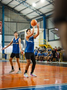 Em quadra, um estudante uniformizado da UFF realiza um arremesso no basquete, enquanto um companheiro observa a jogada. Ao fundo, outros atletas e espectadores acompanham a partida sentados no banco de reservas.