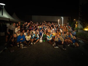 Grupo de estudantes da Atlética Finanças UFF posa para uma fotografia coletiva em área externa de um complexo esportivo durante a noite. Os participantes sorriem para a câmera, alguns segurando instrumentos da bateria da torcida e faixas da atlética, em clima de confraternização.