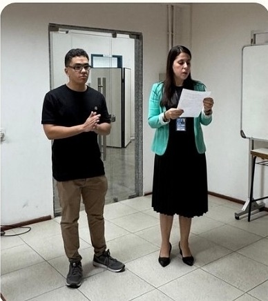 Em uma sala interna com paredes claras e piso de cerâmica, duas pessoas estão em pé lado a lado. À direita, uma mulher de cabelos longos escuros veste blazer verde, vestido preto e sapatos pretos, lendo um texto impresso. À esquerda, uma pessoa de camiseta preta, calça bege e tênis cinza observa a apresentação. Ao fundo, há uma porta aberta de vidro, um quadro branco móvel e parte do mobiliário da sala. O ambiente sugere uma atividade acadêmica ou institucional.