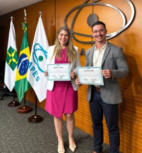 Fotografia em ambiente institucional da CAPES. Duas pessoas estão em pé lado a lado, sorrindo para a câmera e segurando certificados de posse. À esquerda, uma mulher com cabelos longos claros veste blazer branco sobre vestido rosa. À direita, um homem de óculos usa blazer cinza, camisa clara e calça escura. Ao fundo, há painéis de madeira, o logotipo da CAPES e três bandeiras em mastros, incluindo a bandeira do Brasil. A imagem registra um momento formal de celebração e reconhecimento profissional.