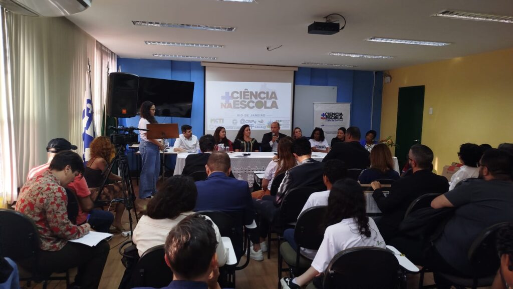 Fotografia de um evento realizado em uma sala de reuniões. Em primeiro plano, dezenas de pessoas estão sentadas em cadeiras voltadas para uma mesa de autoridades posicionada ao fundo. À esquerda, uma mulher está em pé junto a um púlpito, enquanto uma câmera em tripé registra a atividade. Na mesa principal, representantes de instituições participam de uma mesa-redonda; um dos participantes fala ao microfone. Ao centro, uma tela de projeção exibe a identidade visual do programa “Mais Ciência na Escola”, acompanhada de banners do projeto. O ambiente possui paredes em tons de azul e amarelo, iluminação fluorescente e grandes janelas laterais com cortinas claras. O público acompanha atentamente a apresentação.