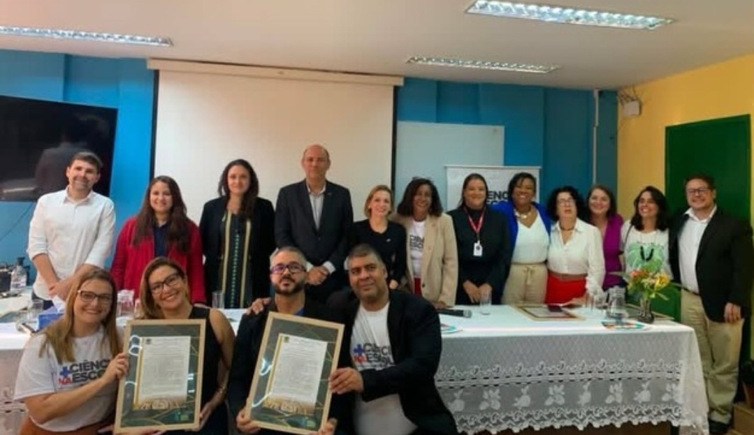 Fotografia registrada em ambiente interno durante uma cerimônia institucional. Cerca de quinze pessoas posam para a foto, distribuídas em duas fileiras diante de uma mesa coberta por toalha branca. Na primeira fila, quatro participantes estão sentados ou ajoelhados, e dois deles exibem quadros com certificados. Ao fundo, autoridades e representantes de instituições permanecem em pé, sorrindo para a câmera. O espaço possui paredes coloridas em tons de azul e amarelo, tela de projeção ao centro e iluminação fluorescente no teto. Alguns participantes usam camisetas com a identificação do programa “Mais Ciência na Escola”.