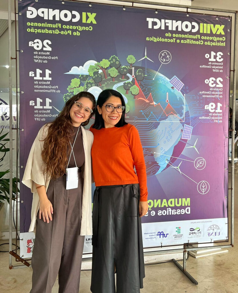 Duas mulheres posam sorrindo diante de um painel de evento acadêmico sobre pós-graduação e inovação científica e tecnológica. À esquerda, uma mulher de cabelos longos ondulados usa óculos, macacão marrom e um casaco claro, com credencial pendurada no pescoço. À direita, uma mulher de cabelos pretos lisos e óculos veste blusa laranja e saia preta. Ao fundo, o painel exibe ilustrações relacionadas à sustentabilidade, tecnologia e meio ambiente, além de informações sobre o congresso. O ambiente é interno e bem iluminado.
