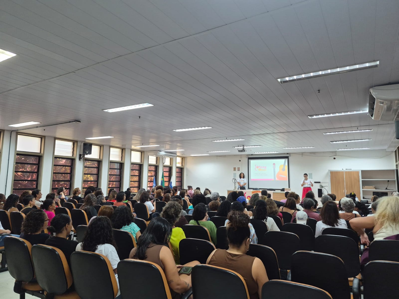 Vista do fundo do auditório durante uma apresentação. À frente, duas mulheres estão no palco, ao lado de uma tela de projeção que exibe a identidade visual da Oficina das Minas. O auditório está cheio, com participantes sentadas em fileiras de cadeiras voltadas para as palestrantes. O ambiente é amplo, iluminado por luz natural e artificial, com janelas ao longo da lateral esquerda.