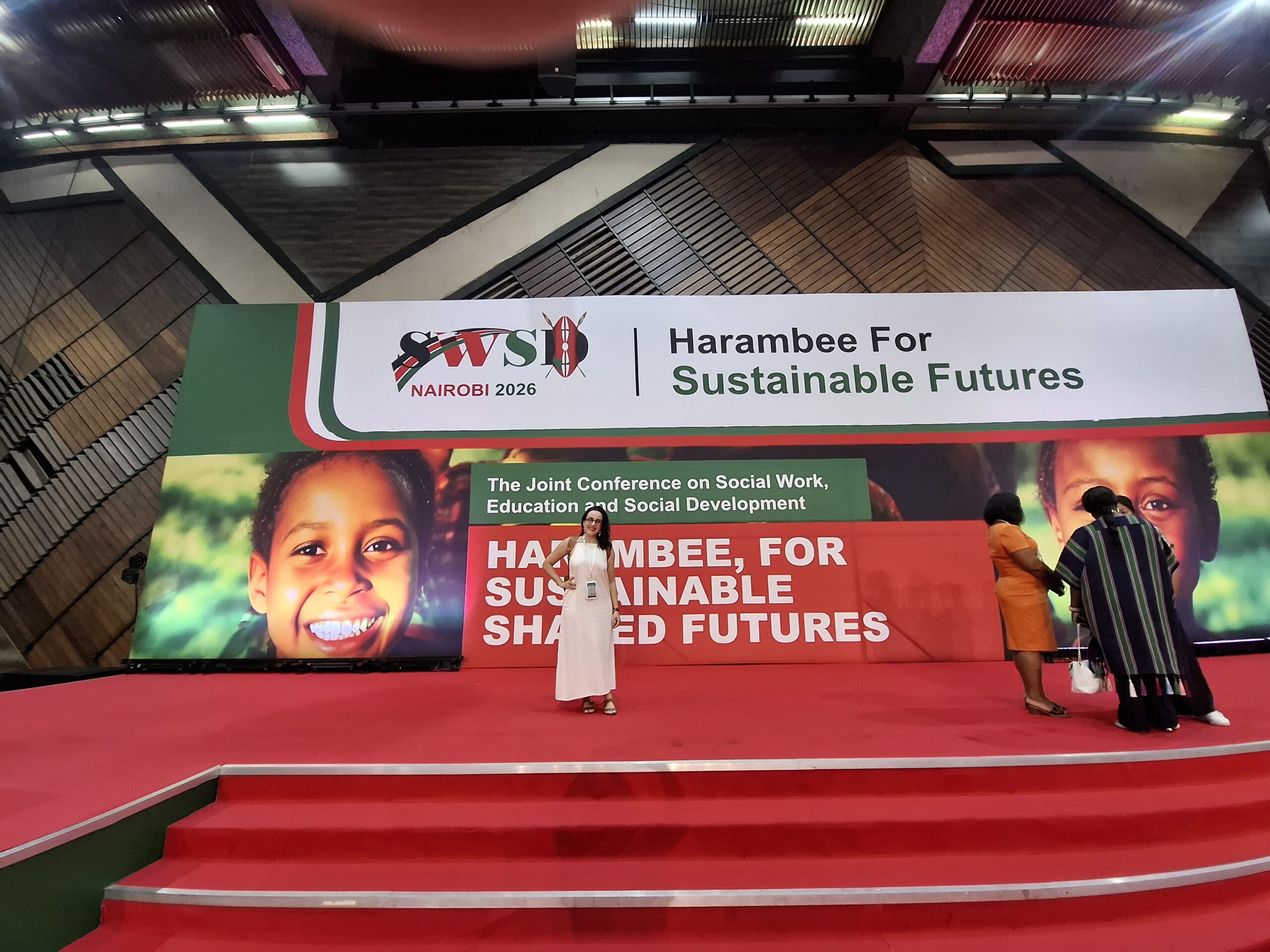 A imagem mostra uma mulher em pé no centro de um palco com carpete vermelho e uma escada que leva ao público. Atrás dela, há um grande banner com o logo da SWSD Nairobi 2026 e a frase "Harambee For Sustainable Shared Futures" escrita em letras brancas sobre um fundo verde e vermelho. O banner também apresenta fotos de crianças negras sorrindo. À direita da mulher, há duas outras mulheres em pé no palco. A mulher à esquerda veste um vestido laranja e a mulher à direita veste um vestido listrado