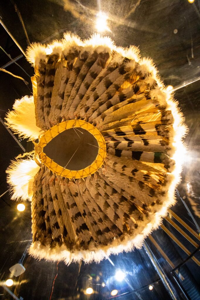 No centro da exposição está o txamatxama, artefato plumário de profundo significado cultural, espiritual e político para os povos Katxuyana e Kahyana. Associado à identidade, à transmissão de conhecimentos e a práticas tradicionais, o objeto também representa um importante elemento de conexão entre pessoas, territórios, memórias e diferentes mundos. Crédito: Woltaire Masaki/MPEG